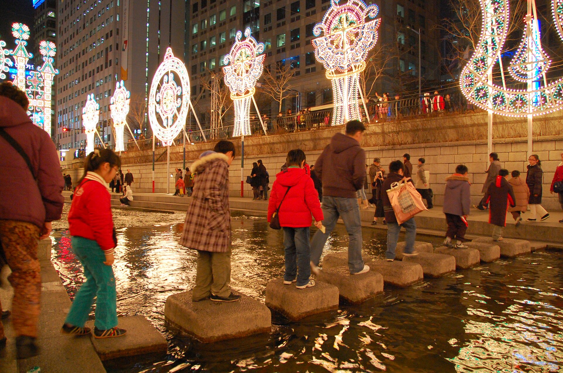 Families crossing stepping stones under winter lights on Cheonggyecheon Stream in Seoul